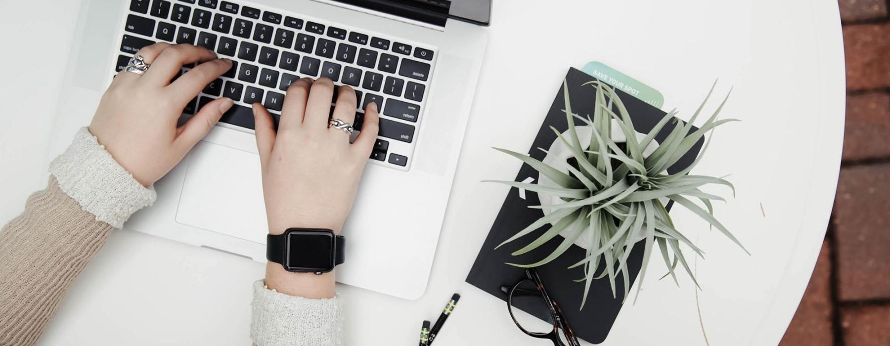 overhead shot of woman working on a laptop at a round table with pencils, her cell phone, glasses, notebook and a potted plant